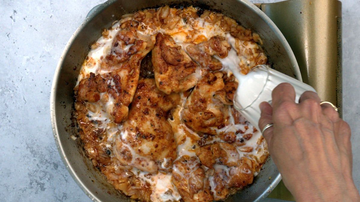 A hand pours coconut milk over browned chicken pieces and sautéed shallots in a skillet, creating a rich sauce. The dish is being prepared on a light gray countertop.