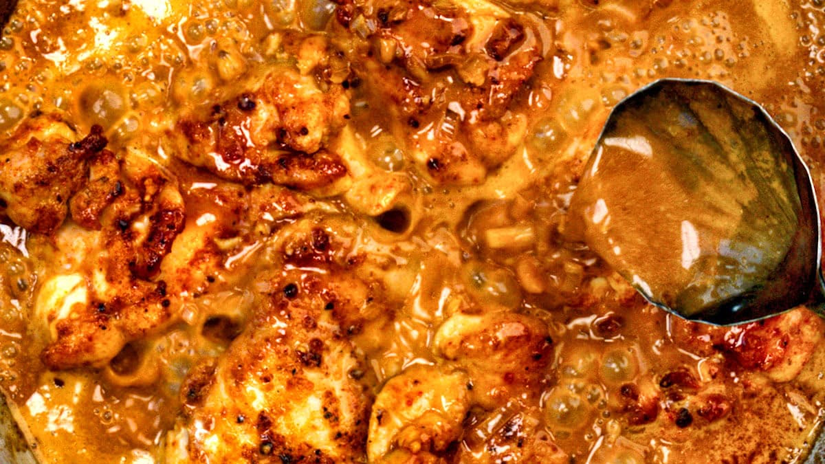 Golden brown chicken pieces simmer in a bubbling, creamy sauce as a metal spoon dips into the mixture, capturing the rich, flavorful texture of the dish.