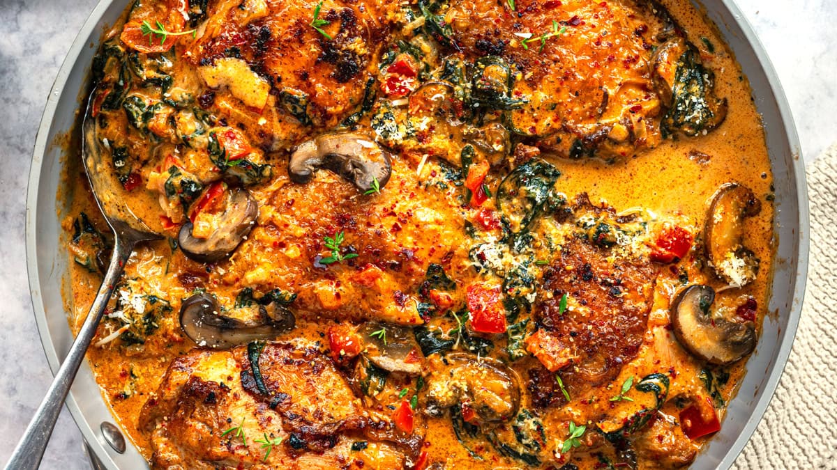 A skillet filled with creamy chicken paprika Tuscan-style, featuring golden-brown chicken thighs, mushrooms, spinach, diced red peppers, and a rich, orange-hued sauce, with a spoon resting in the dish.