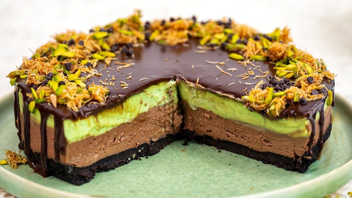 A layered chocolate and green cake with a cookie crust, topped with chocolate glaze, chopped pistachios, and toasted flakes, sits on a green plate with a slice removed to show its creamy texture.