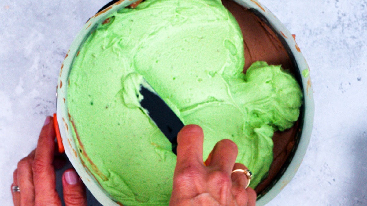 A person spreads light green pistachio cheesecake filling over a chocolate base in a round pan, using a spatula. The background is a light, textured surface.