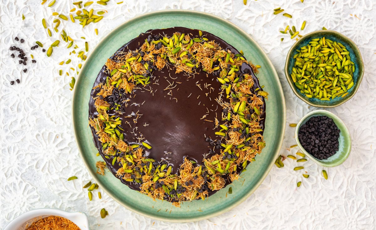 A round Dubai chocolate cheesecake topped with chopped pistachios and shredded phyllo on a green plate, surrounded by small bowls of sliced pistachios and chocolate chips on a white lace tablecloth.