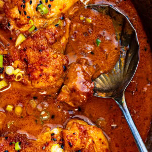 Close-up of gochujang chicken thighs in a rich, reddish-brown sauce, garnished with chopped green onions and black sesame seeds. A metal spoon is partially submerged in the sauce beside the chicken pieces.