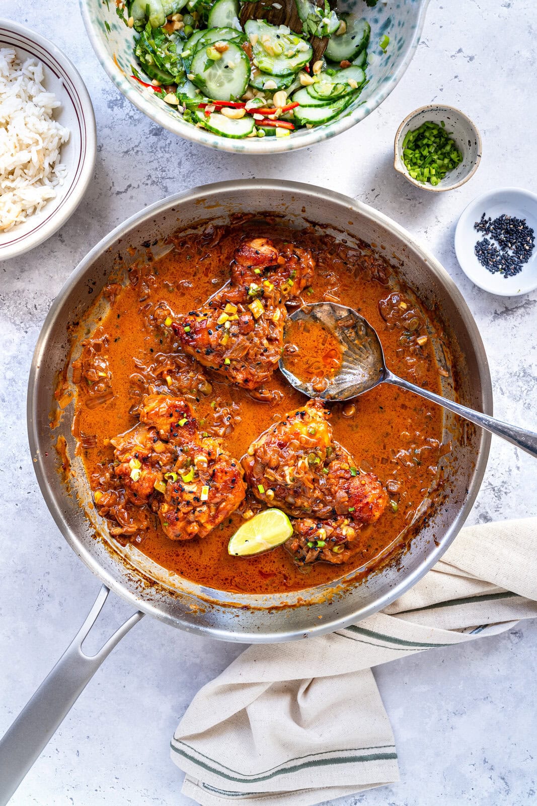A skillet with four pieces of chicken thighs in a red, saucy curry garnished with corn and herbs, a spoon resting in the pan. Nearby are bowls of white rice, cucumber salad, chopped green onions, and black sesame seeds.