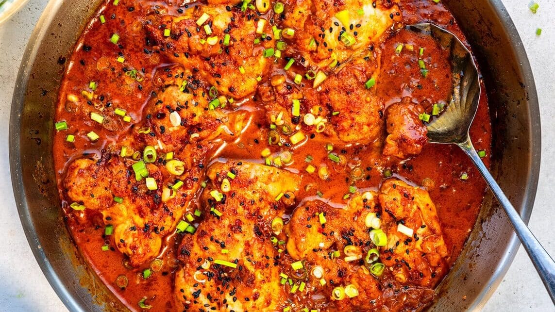 A pan of chicken simmering in a rich, red sauce, garnished with chopped green onions and black sesame seeds. A spoon rests in the pan. Nearby are a bowl of rice, a salad, a halved lime, and a small dish of black seeds.