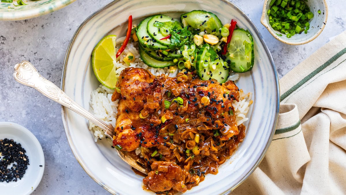 A plate with rice, glazed chicken topped with onions and herbs, cucumber salad with peanuts and chili, a lime wedge, and a sprinkle of black sesame seeds, with small bowls of garnishes on the side.