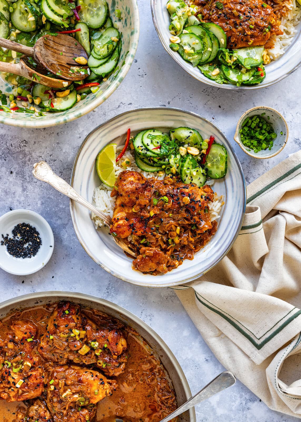 Overhead view of plates with chicken in a rich sauce served over rice, garnished with green onions and chilies, accompanied by cucumber salad. A skillet with more chicken and a bowl of chives are also visible.