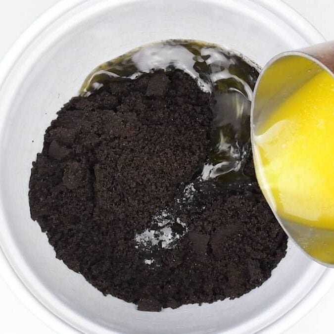 Crushed chocolate cookies in a white bowl with melted butter being poured over them from a metal cup, likely for making a dessert crust.