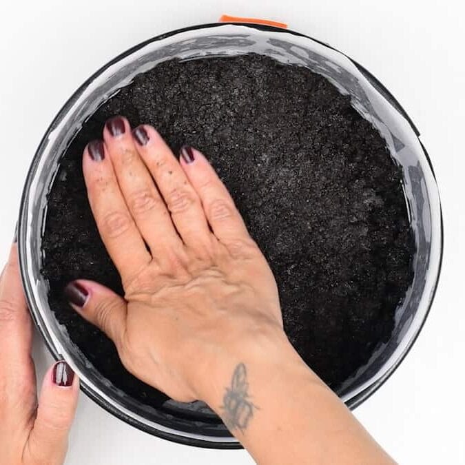 A hand with dark nail polish and a bee tattoo presses down a layer of dark crumb crust into a round pan lined with parchment paper.