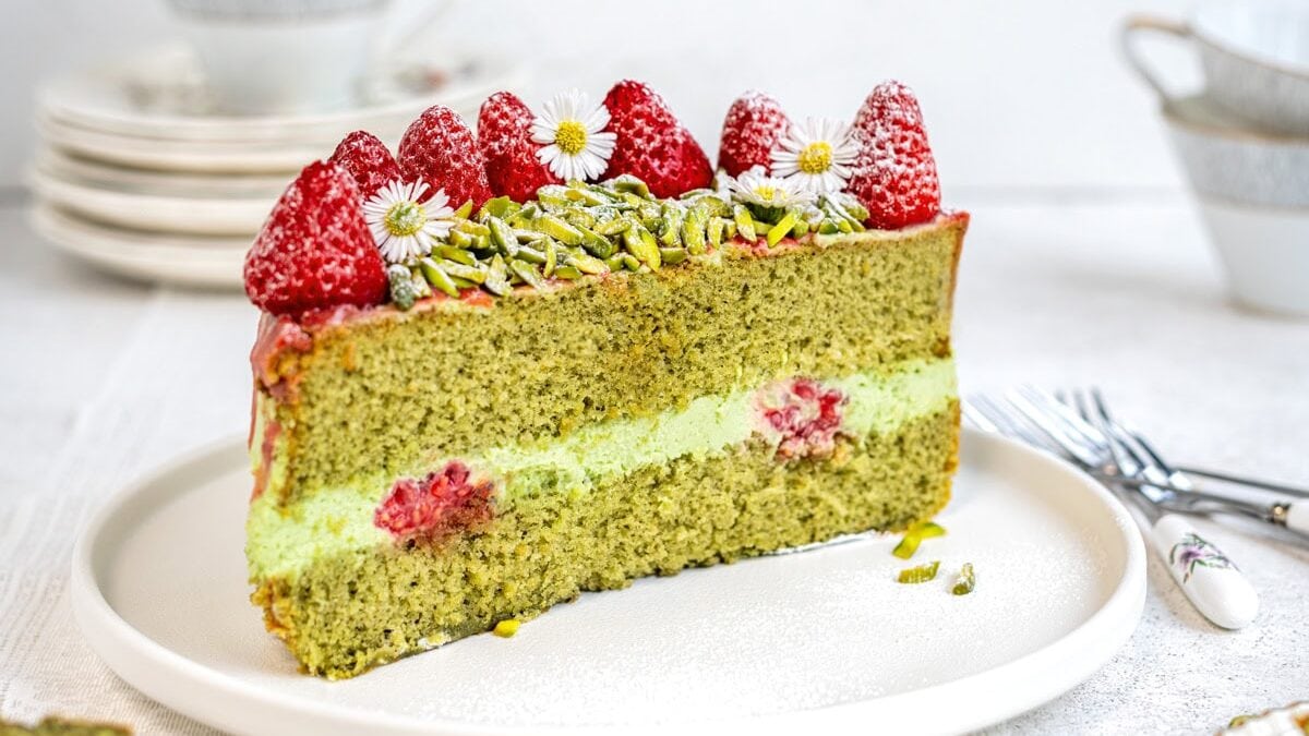 A slice of green tea cake with a light green filling, decorated with strawberries, chopped pistachios, and small white flowers on top, served on a white plate with stacked dishes and cutlery in the background.