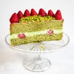 A slice of green matcha chiffon cake with cream and raspberry filling, topped with whole strawberries and sliced pistachios, displayed on a clear glass cake stand against a white background.
