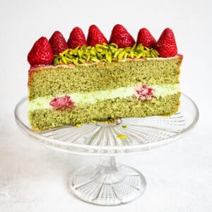 A slice of green matcha chiffon cake with cream and raspberry filling, topped with whole strawberries and sliced pistachios, displayed on a clear glass cake stand against a white background.
