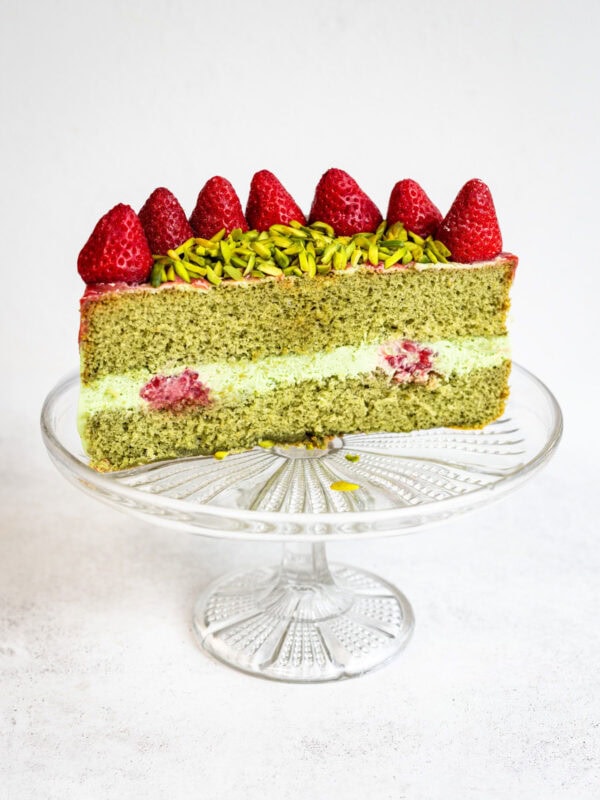 A slice of green matcha chiffon cake with cream and raspberry filling, topped with whole strawberries and sliced pistachios, displayed on a clear glass cake stand against a white background.