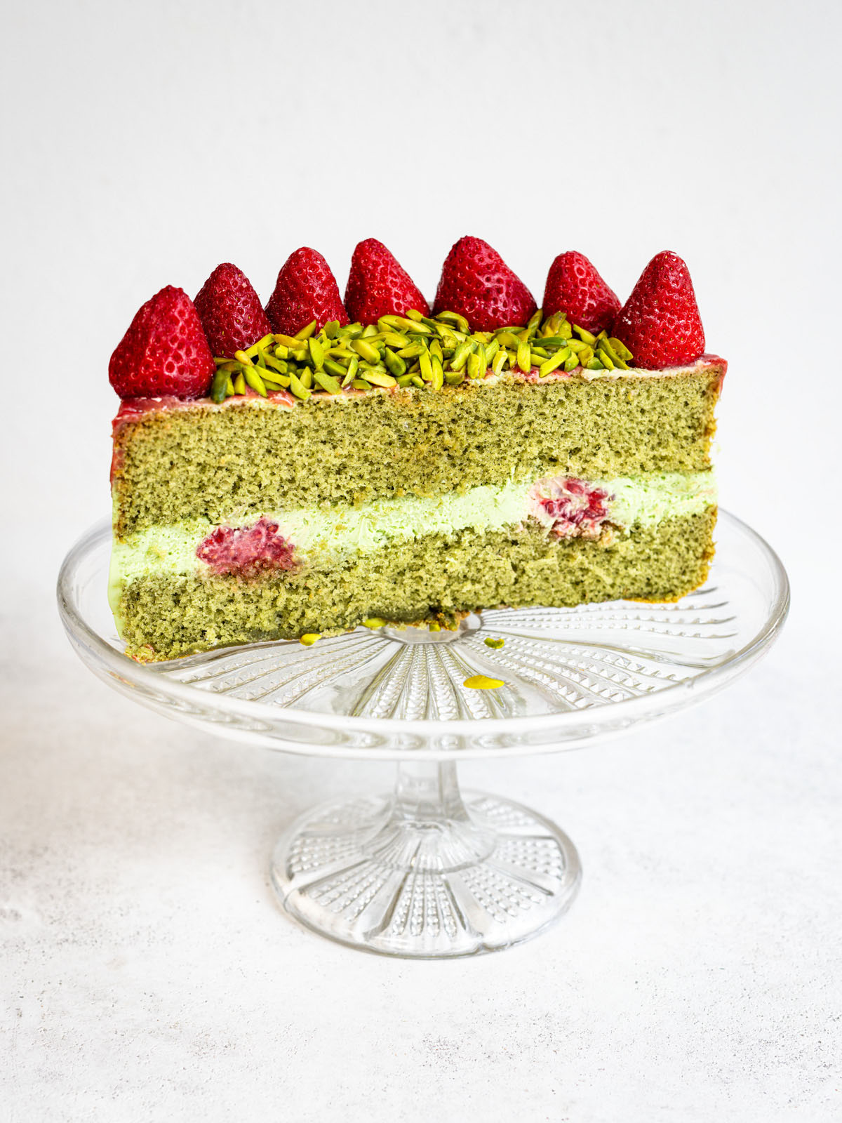 A slice of green sponge cake with a creamy filling, raspberries inside, topped with sliced pistachios and whole strawberries, displayed on a glass cake stand against a white background.