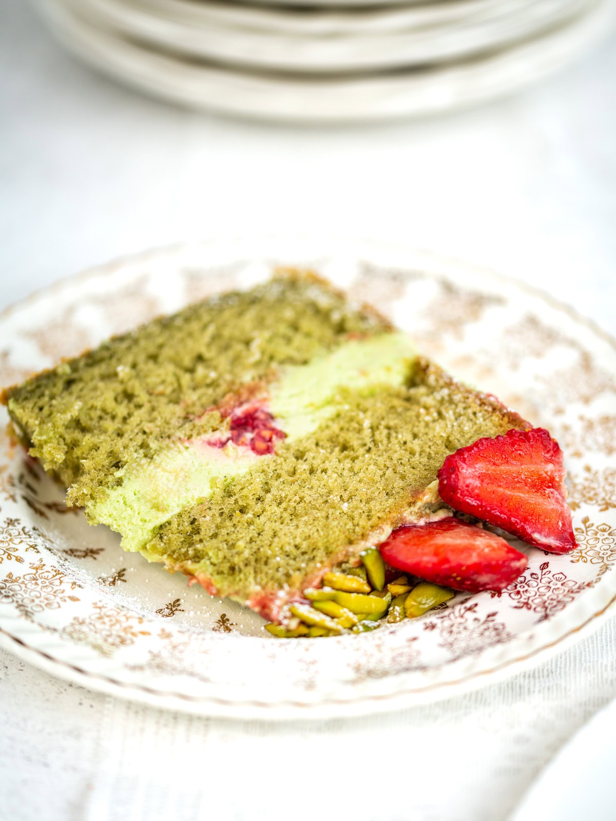 A slice of green matcha cake with a creamy layer and a raspberry center, served on a decorative plate with strawberries and chopped pistachios on the side.