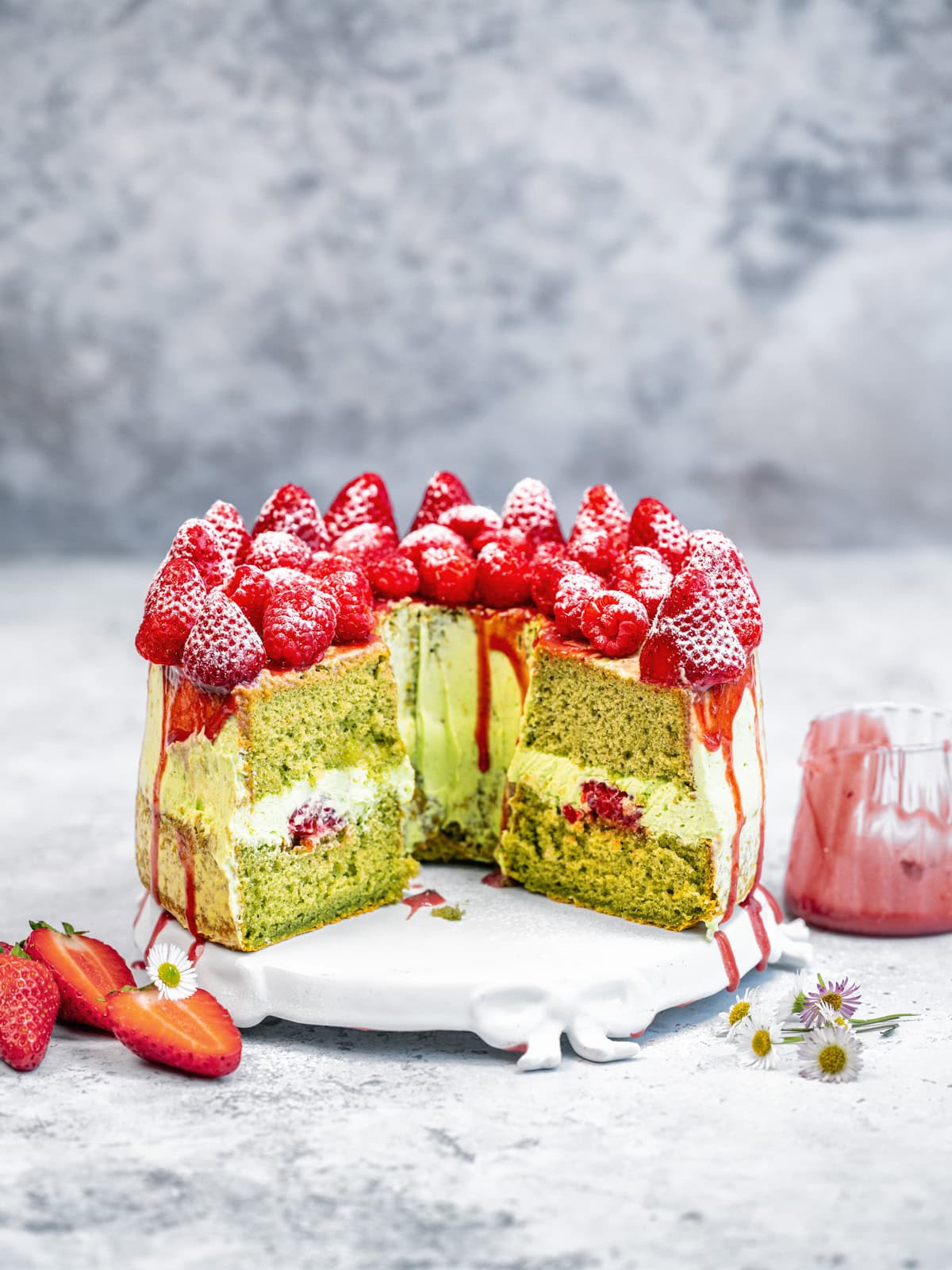 A tall matcha cake with a slice cut out, filled with cream and strawberries, topped with whole strawberries and powdered sugar, sits on a white stand; strawberries and flowers are scattered nearby.