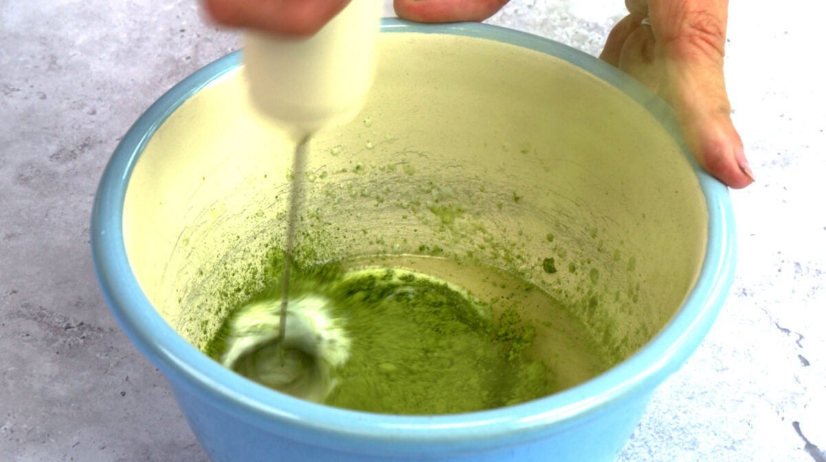 A hand-held electric whisk is mixing green matcha powder with buttermilk in a light blue bowl on a light gray surface.