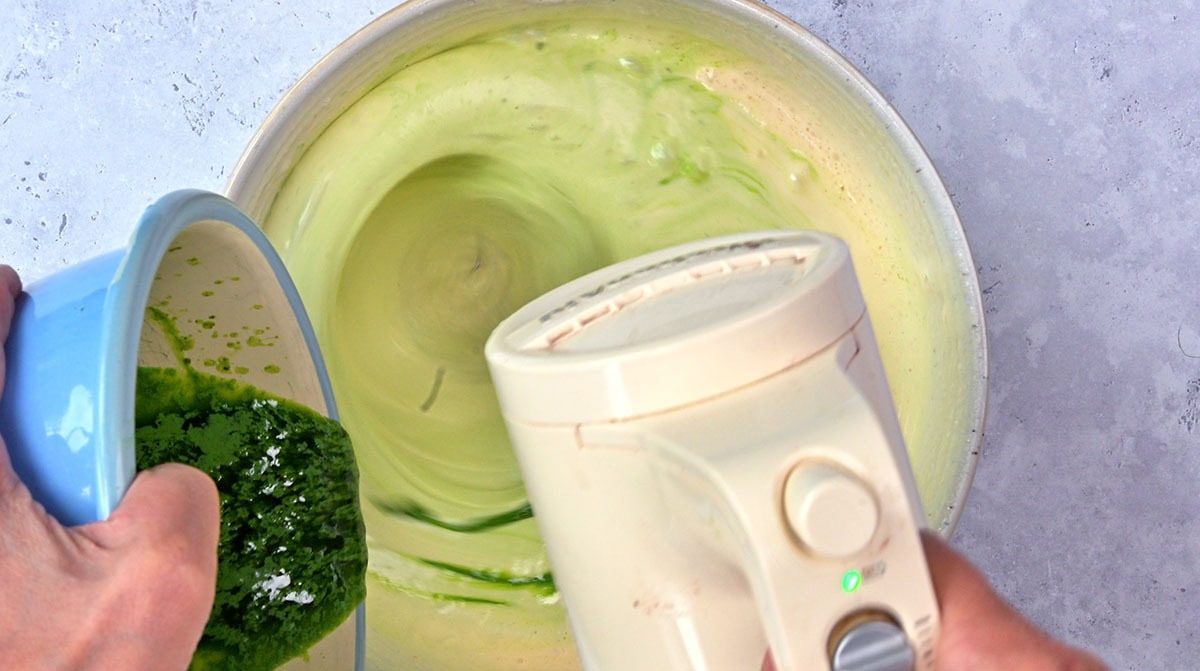 A person uses a handheld electric mixer to blend ingredients in a bowl while pouring a green mixture from a blue bowl into the batter.
