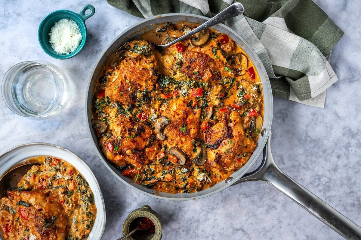 A skillet filled with creamy chicken thighs, mushrooms, spinach, and red peppers in a rich sauce. A serving dish, bowl of grated cheese, glass of water, and green-and-white cloth napkin sit nearby on a marble surface.