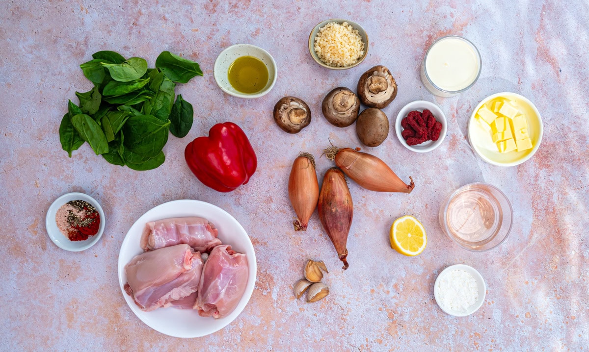 An assortment of ingredients on a pink surface, including raw chicken, spinach, red bell pepper, shallots, mushrooms, butter, grated cheese, olive oil, cream, tomato paste, garlic, lemon, flour, and various spices in small bowls.