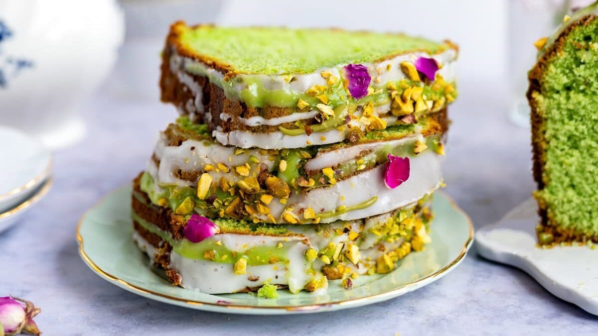 Three slices of vibrant green pistachio loaf cake with white icing, chopped pistachios, and rose petals are stacked on a plate. A teapot, stacked cups, and a vase of flowers are in the background.