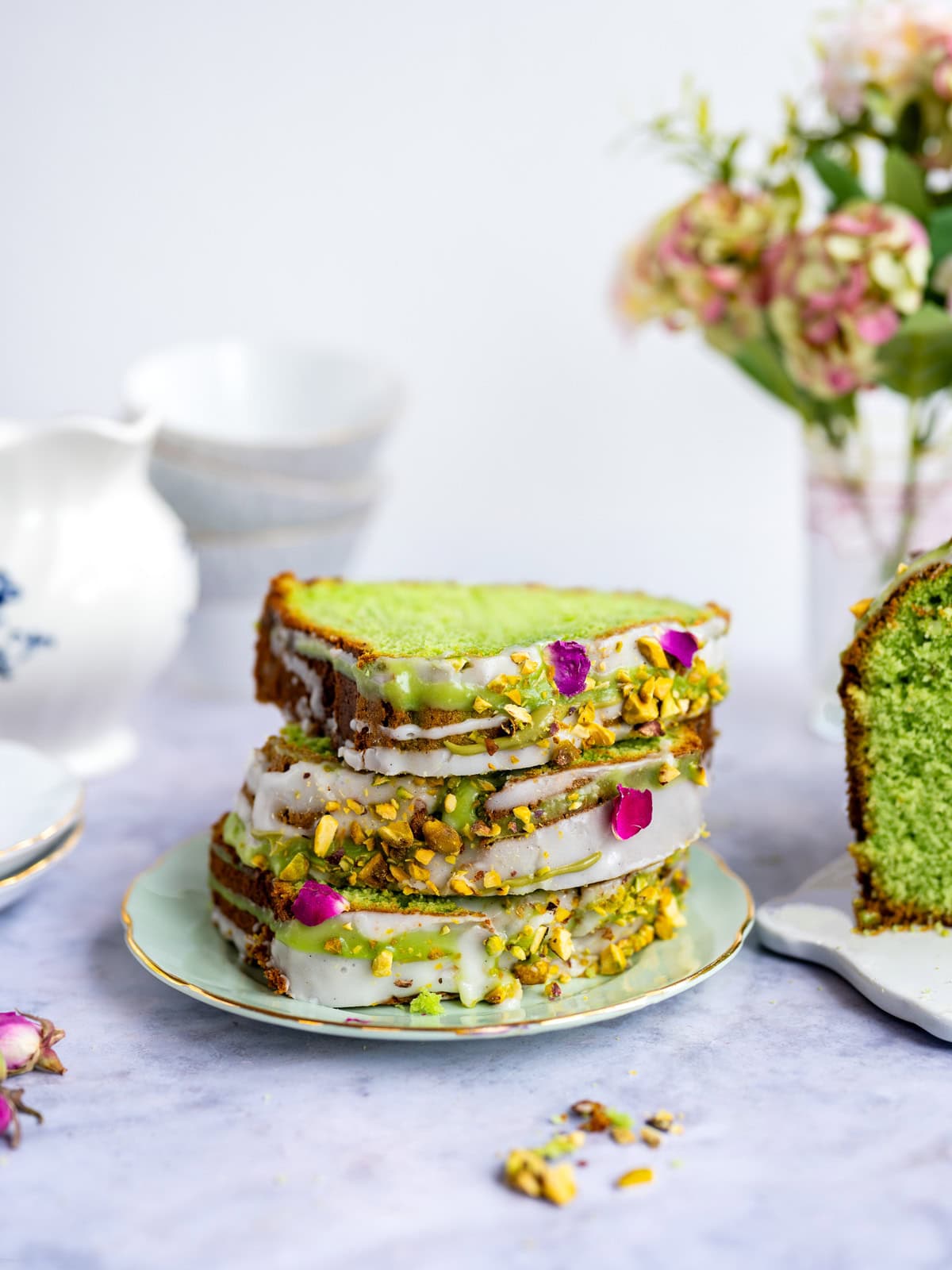 Three slices of vibrant green pistachio loaf cake with white icing, chopped pistachios, and rose petals are stacked on a plate. A teapot, stacked cups, and a vase of flowers are in the background.