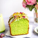 A pistachio bread loaf cake with white icing, topped with chopped pistachios and pink flower petals, sits on a white tray. In the background, a vase of pink flowers is softly blurred.
