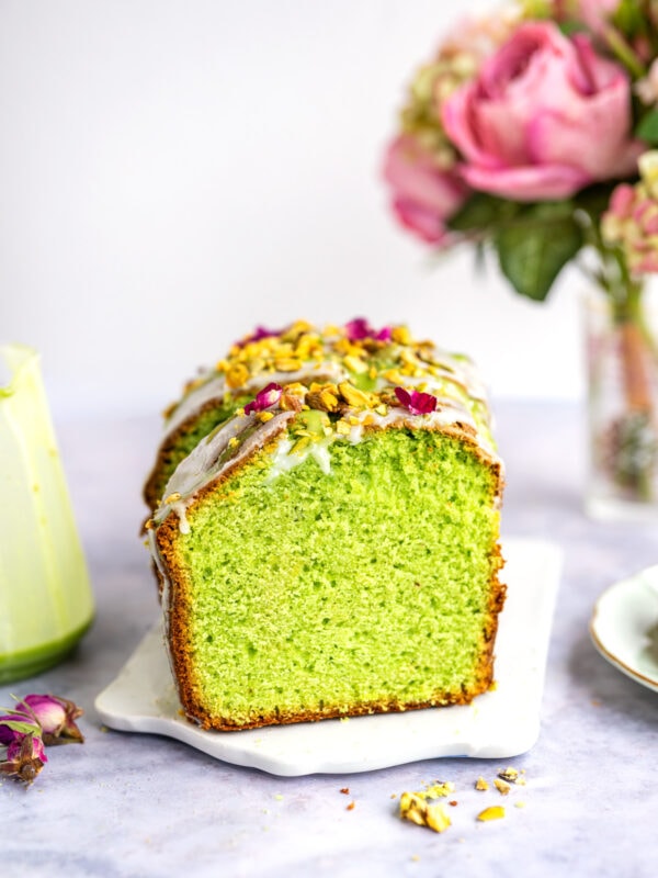 A pistachio bread loaf cake with white icing, topped with chopped pistachios and pink flower petals, sits on a white tray. In the background, a vase of pink flowers is softly blurred.