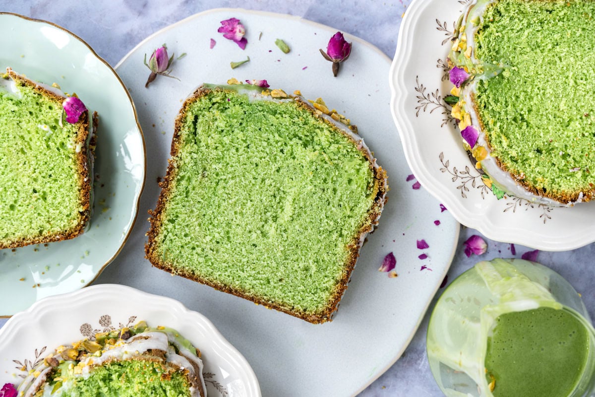A thick slice of vibrant green pistachio cake with glaze, served on a decorative plate, surrounded by more slices on plates and scattered pink flower petals. A green drink is nearby.