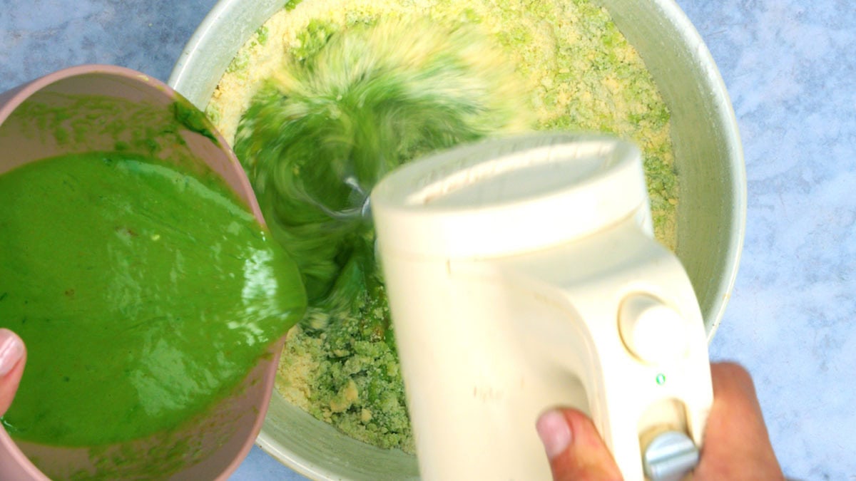 A person pours thick green liquid ingredients from a bowl into a mixing bowl while using a hand mixer to blend ingredients, creating a swirling motion in the mixture.