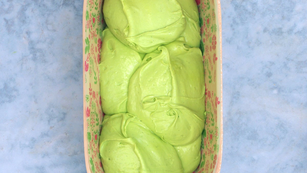 A rectangular floral dish filled with bright green, pistachio loaf cake batter placed on a blue marbled surface.