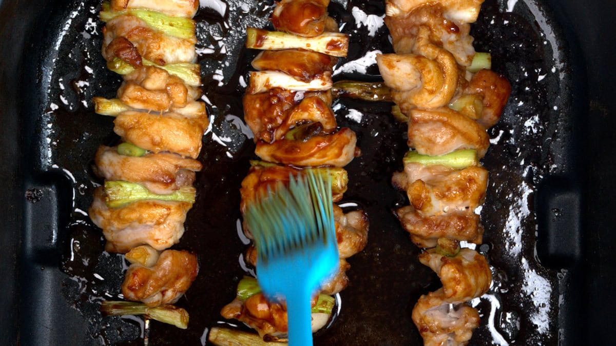 Three chicken and vegetable skewers are being brushed with a glossy brown sauce using a blue brush, inside a dark cooking pan.