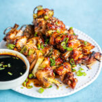 Air fryer chicken yakitori skewers garnished with chopped green onions, served on a white plate with a small bowl of dark dipping sauce on the side, set against a light blue background.