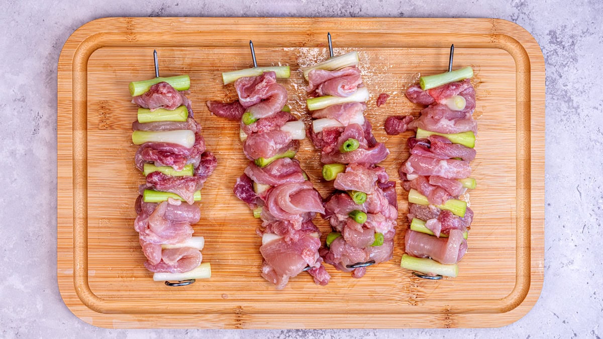 Four metal skewers with alternating pieces of raw meat and green onions are arranged on a wooden cutting board, ready for grilling. The board sits on a light-colored surface.