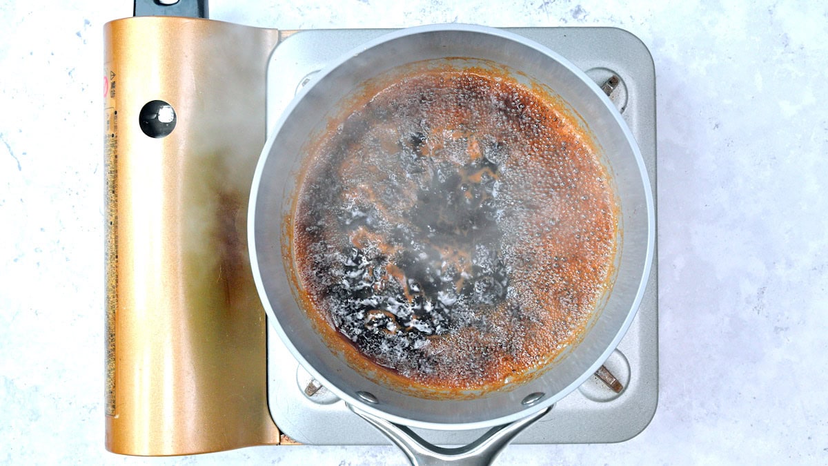 A pot filled with yakitori sauce is cooking on a portable electric burner against a light, textured background. Steam rises from the bubbling surface.