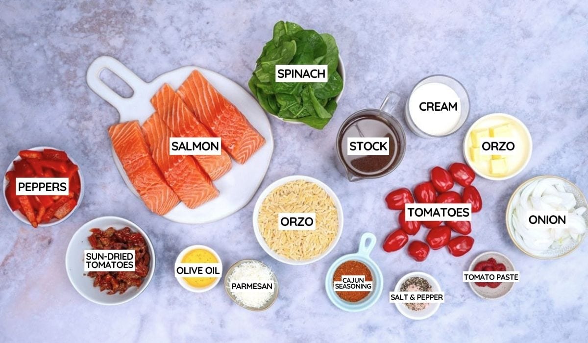 Ingredients for a creamy Cajun Salmon Orzo dish are arranged on a table, featuring salmon fillets, spinach, orzo pasta, cream, stock, tomatoes, onion, peppers, sun-dried tomatoes, olive oil, parmesan, and seasonings.