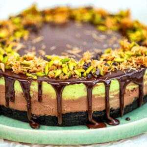 A close-up of a layered cheesecake with chocolate ganache dripping down the sides, topped with chopped pistachios and toasted shredded coconut, sitting on a light green plate.