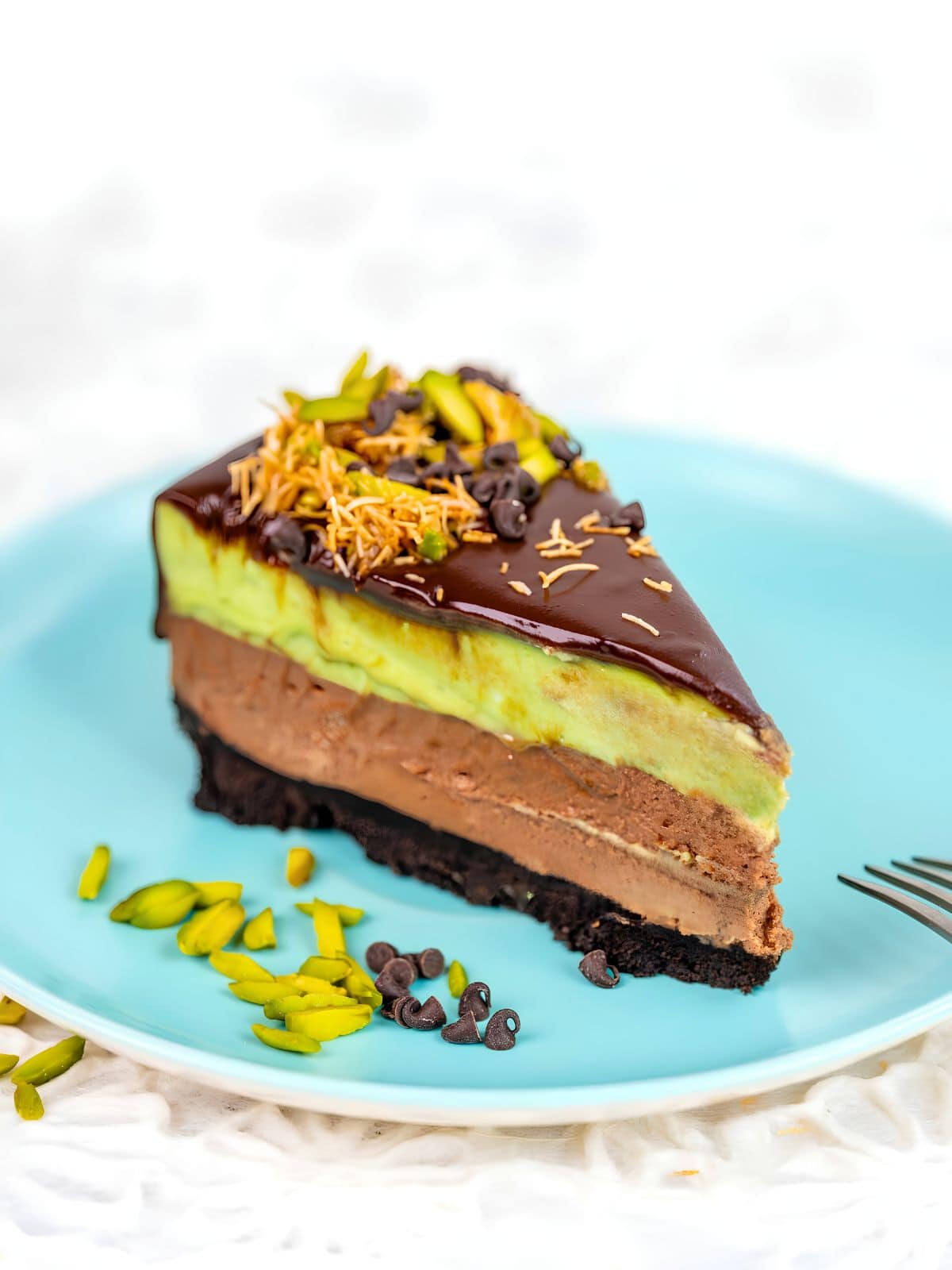 A slice of layered chocolate and pistachio cake with chocolate ganache, reminiscent of a Dubai chocolate cheesecake, sits on a blue plate with a fork and scattered pistachios and chips on the side.