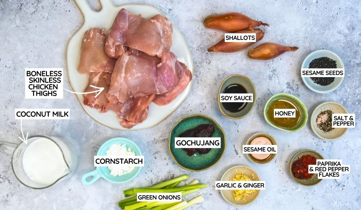 A flat lay of labeled ingredients for a chicken dish, including boneless skinless chicken thighs, coconut milk, shallots, soy sauce, honey, sesame oil, gochujang, cornstarch, green onions, garlic & ginger, sesame seeds, salt & pepper, and paprika & red pepper flakes.