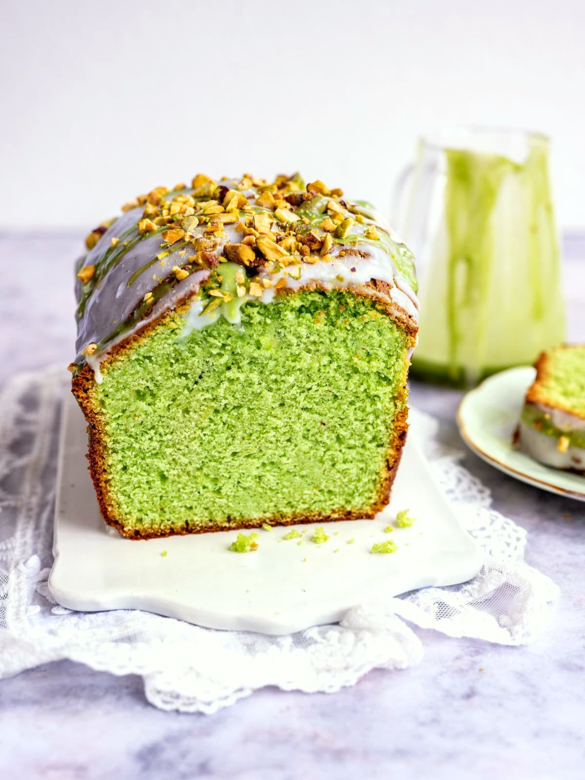 A loaf of pistachio loaf cake with white icing and chopped pistachios on top sits on a white board. A slice has been cut off, revealing the vibrant green interior. A jar and a plate with a cake slice are in the background.