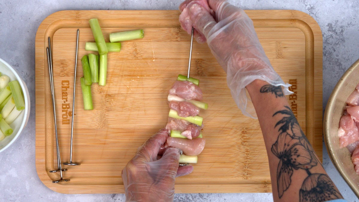 A person wearing gloves threads pieces of raw chicken and green onions onto a metal skewer on a wooden cutting board, with extra skewers and sliced onions nearby.