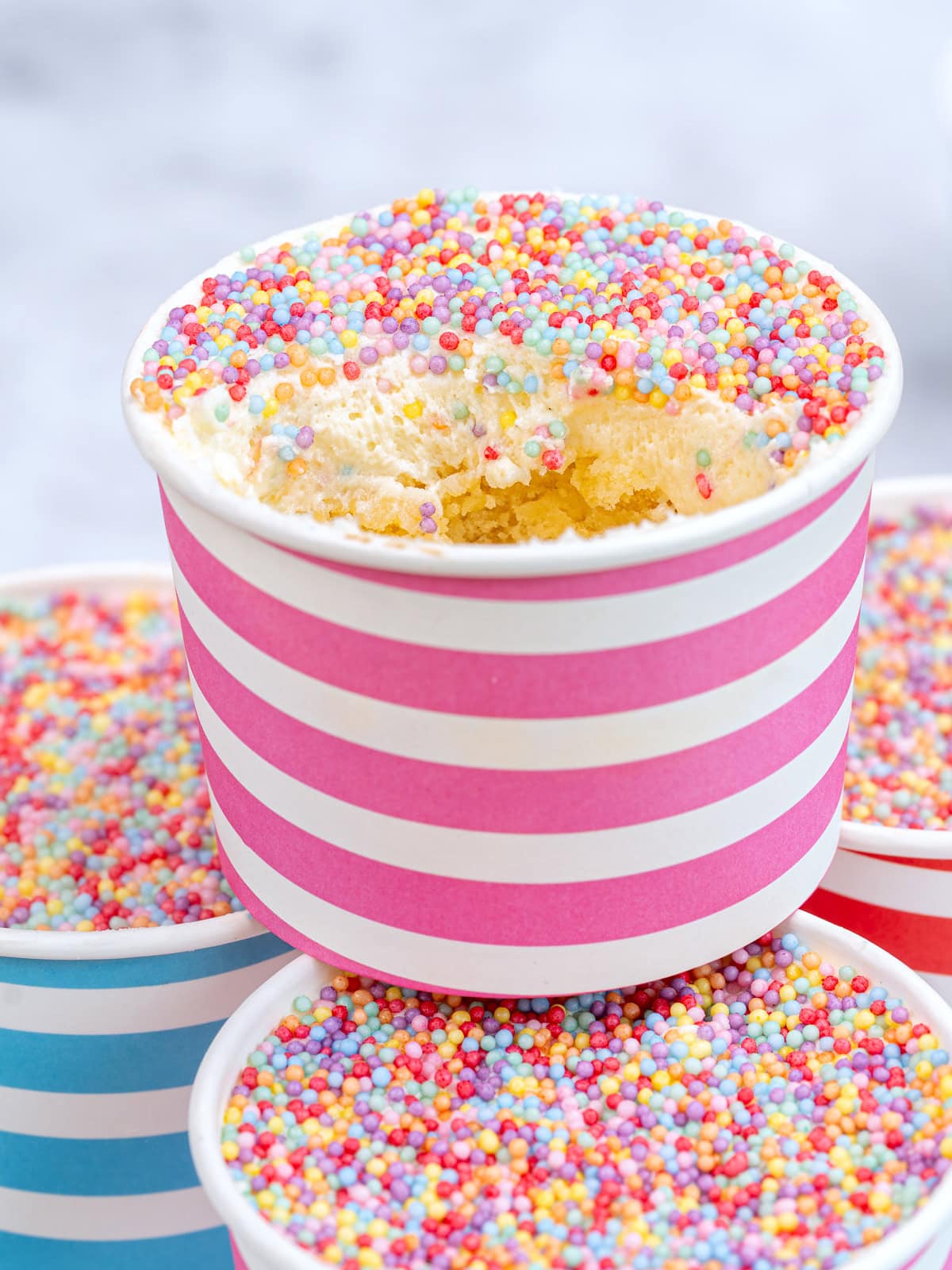 A pink-striped paper cup filled with cake topped with colorful sprinkles, with a bite taken out, sits on top of other sprinkle-topped cups. The background is softly blurred.