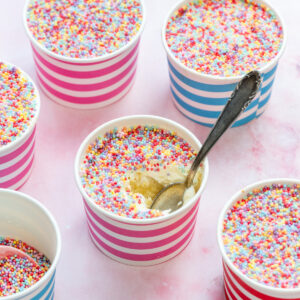 Homemade dot cakes: vanilla cake topped with vanilla buttercream and topped with nonpareil sprinkles, some with a spoon inside. The cups have blue and pink stripes and are arranged on a light pink surface.