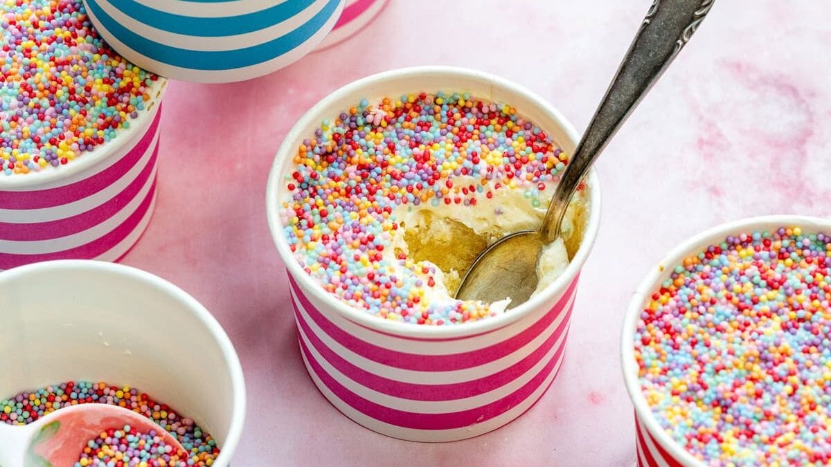Homemade dot cakes: vanilla cake topped with vanilla buttercream and topped with nonpareil sprinkles, some with a spoon inside. The cups have blue and pink stripes and are arranged on a light pink surface.