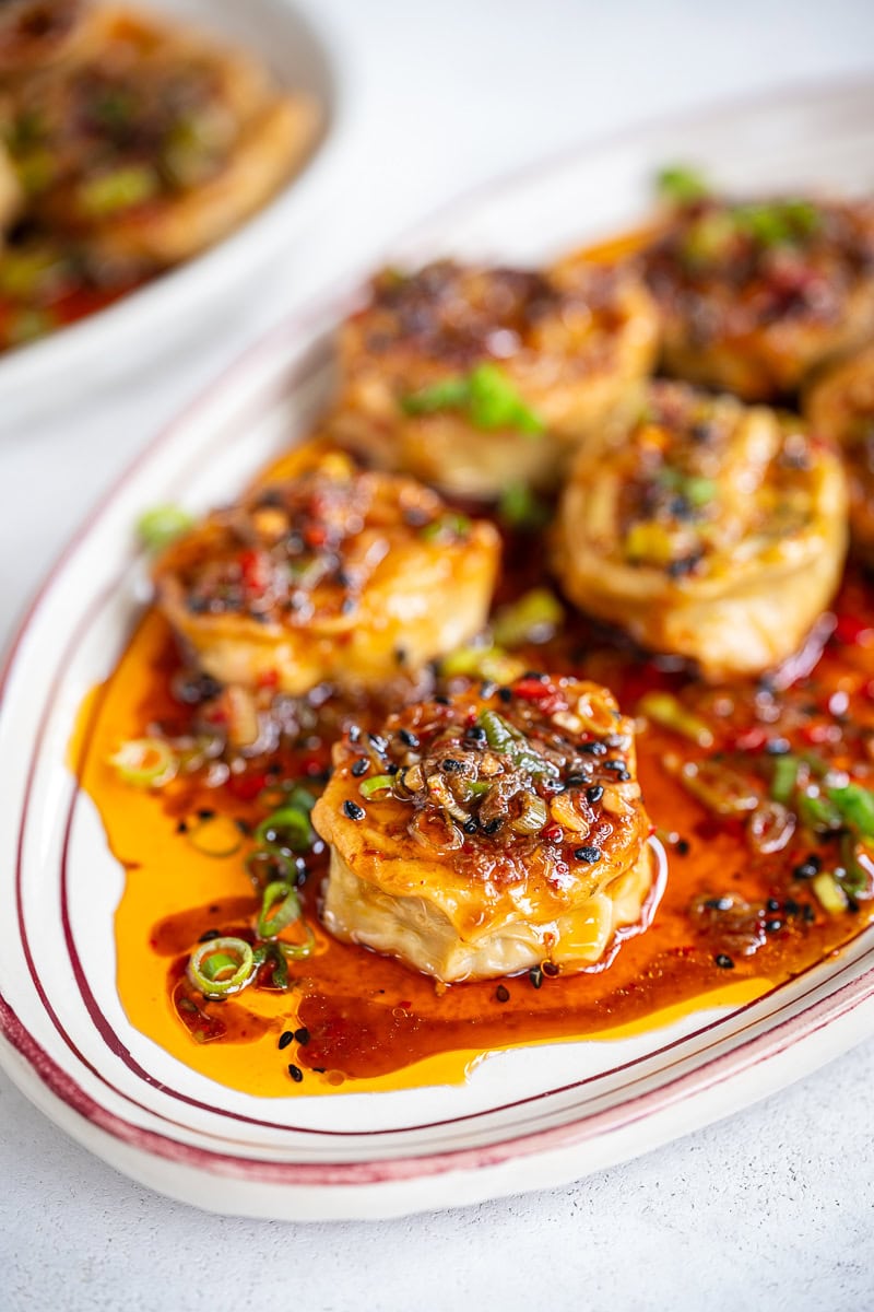 A plate of dumpling rolls topped with a savory sauce, garnished with chopped green onions and sesame seeds, sits on a white dish with red trim. The sauce is glossy and vibrant, pooling around the tofu pieces.