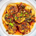 Six pan-fried dumplings topped with chopped green onions and red chilies sit in a white bowl, surrounded by a spicy chili oil sauce with visible pepper flakes and herbs.