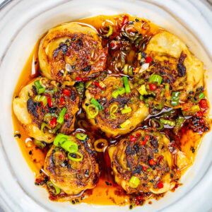 Six pan-fried dumplings topped with chopped green onions and red chilies sit in a white bowl, surrounded by a spicy chili oil sauce with visible pepper flakes and herbs.