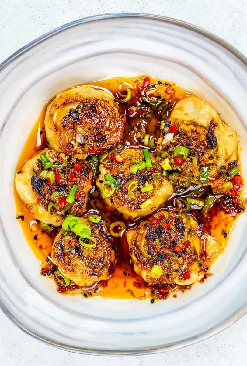 Six pan-fried dumplings topped with chopped green onions and red chilies sit in a white bowl, surrounded by a spicy chili oil sauce with visible pepper flakes and herbs.