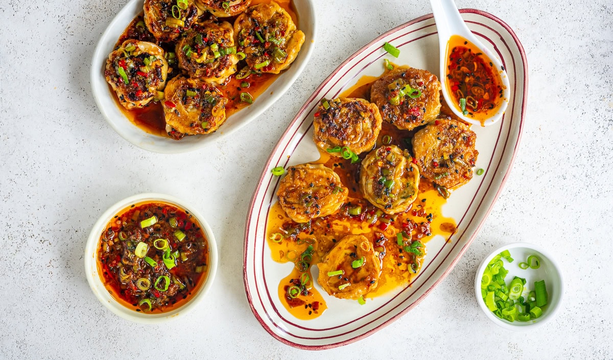 Two plates of pan-fried dumplings topped with chopped green onions and chili oil, with bowls of dipping sauce and sliced green onions, arranged on a white surface.
