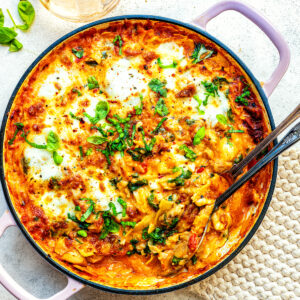 A skillet filled with cheesy baked Marry Me chicken lasagna topped with melted mozzarella and fresh basil, with serving utensils inside. A glass of white wine and a beige textured cloth are beside it on a light surface.